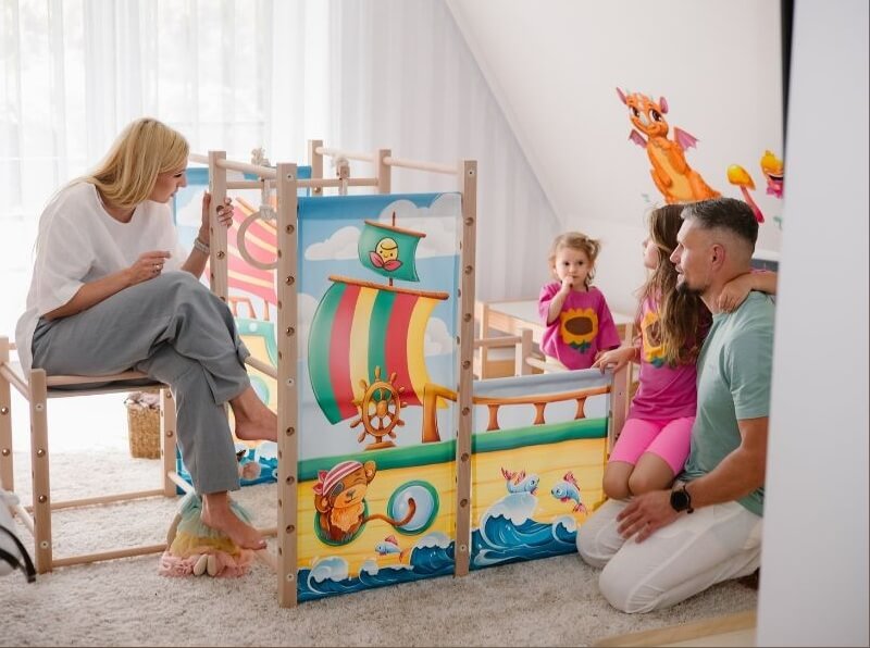 Na zdjęciu widać rodzinę spędzającą wspólnie czas przy domowym, drewnianym placu zabaw ustawionym w jasnym pokoju dziecięcym. Konstrukcja została ozdobiona kolorowymi panelami z ilustracją statku, fal i bajkowych postaci, co nadaje jej charakter pirackiej przygody. Mama siedzi na jednym z elementów zestawu, obserwując dzieci z uśmiechem, natomiast tata klęczy obok, wspierając córkę stojącą przy konstrukcji. W tle widać kolejne bajkowe dekoracje na ścianie, które tworzą przyjazną, kreatywną atmosferę.