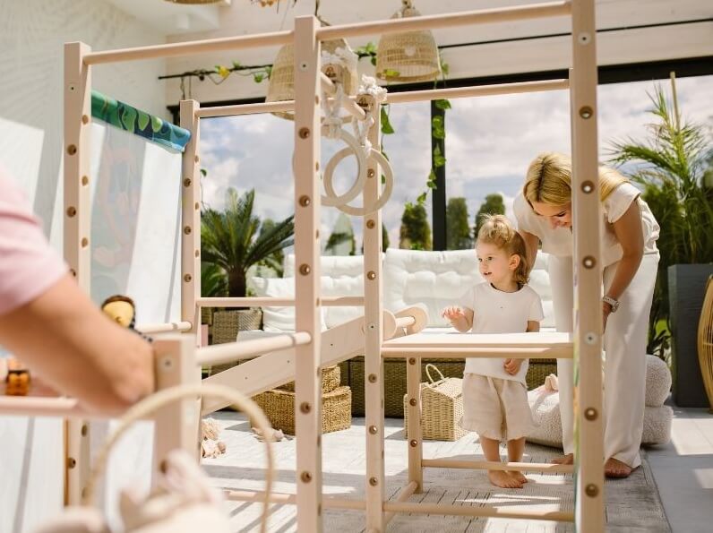 Na zdjęciu widać jasne, przestronne wnętrze z dużymi oknami wychodzącymi na ogród. W centrum stoi drewniany, modułowy plac zabaw z drążkami i kółkami gimnastycznymi. Przy konstrukcji znajduje się małe dziecko, które stoi na platformie i odkrywa kolejne elementy zestawu. Obok pochyla się kobieta, czuwając nad zabawą i wspierając malucha w bezpiecznym korzystaniu z konstrukcji.