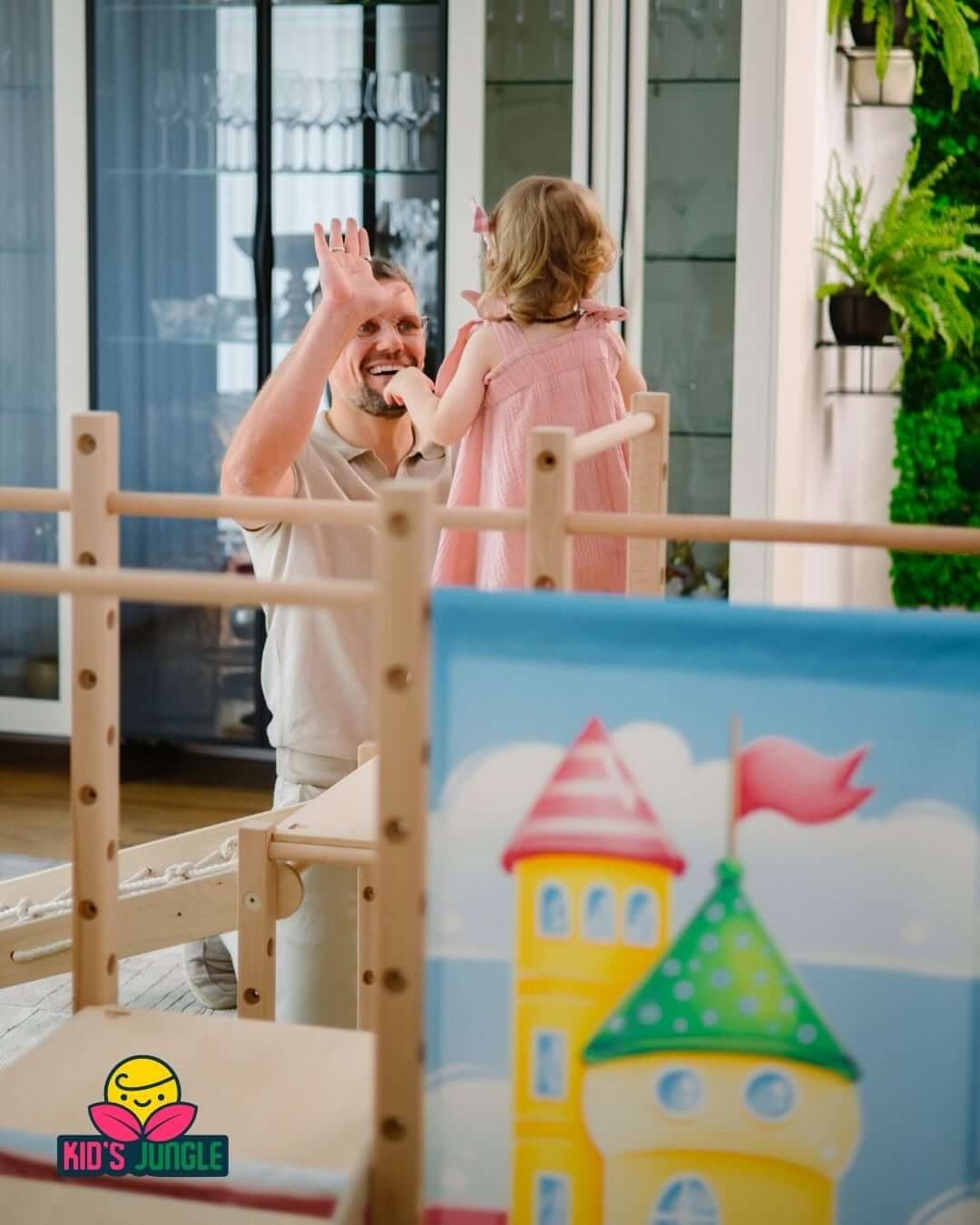 Na zdjęciu widać małe dziecko bawiące się na drewnianym placu zabaw w domu. W tle rodzic z uśmiechem zachęca malucha do zabawy, a kolorowy panel z motywem bajkowego zamku nadaje przestrzeni przyjazny i radosny charakter.