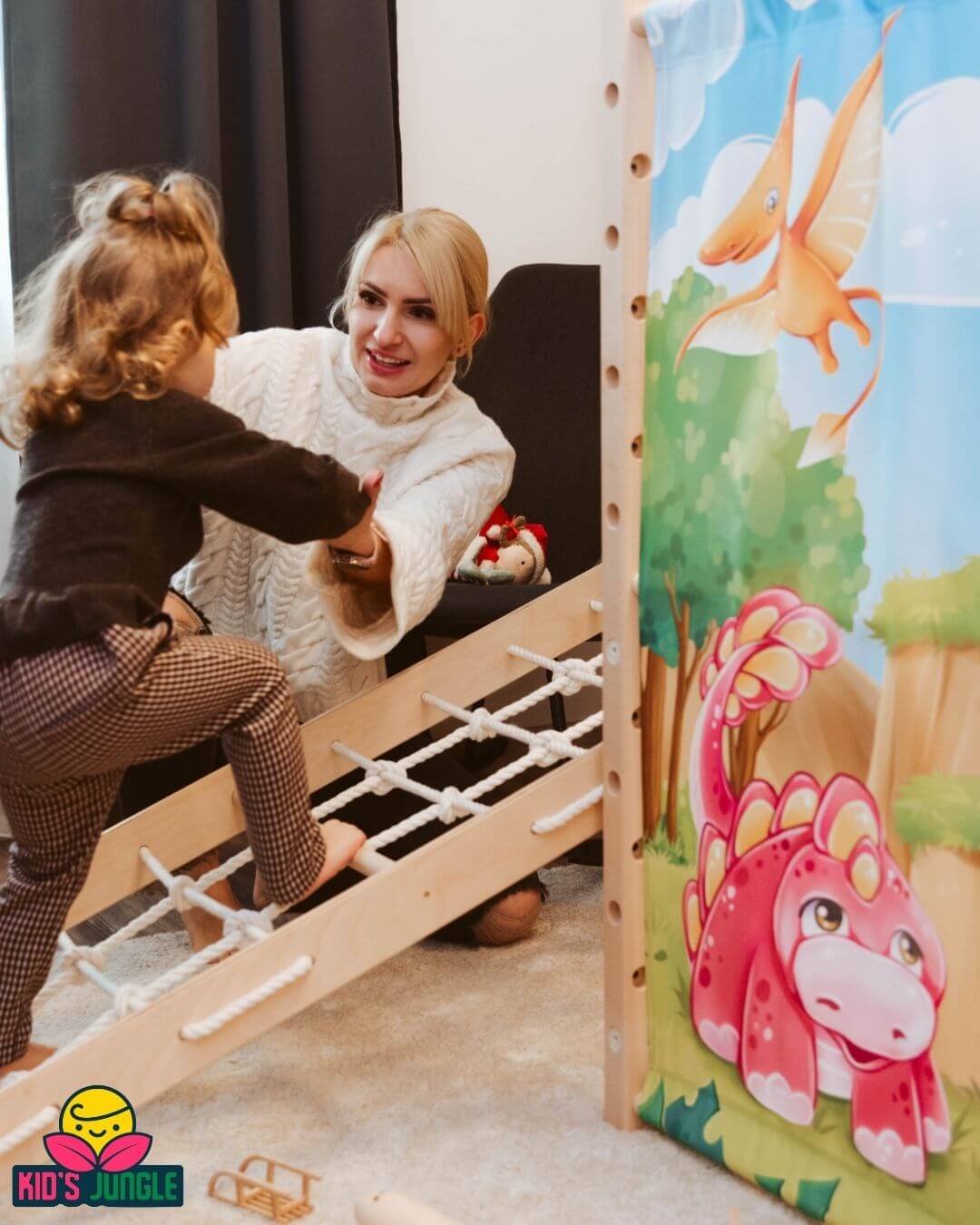 Na zdjęciu widać małe dziecko wspinające się po drewnianej drabince linowej w domowym placu zabaw. Obok klęczy kobieta, która podtrzymuje malucha za ręce i z uśmiechem wspiera go w pokonywaniu kolejnych szczebli. Scena rozgrywa się w przytulnym wnętrzu – konstrukcja stoi na miękkim dywanie, a z boku znajduje się kolorowa ścianka z ilustracjami dinozaurów i bajkowego krajobrazu.