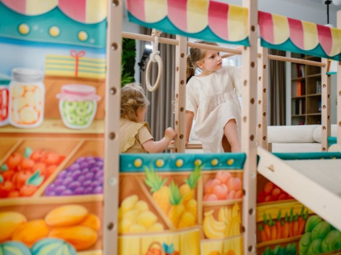 Na zdjęciu widać dwójkę małych dzieci bawiących się na kolorowej, drewnianej konstrukcji. Dzieci są zaangażowane w zabawę — jedno wspina się po drabince, drugie stoi obok, obserwując lub szykując się do ruchu. Konstrukcja zawiera także kółka gimnastyczne, co sugeruje, że łączy funkcje ruchowe z kreatywną zabawą. 